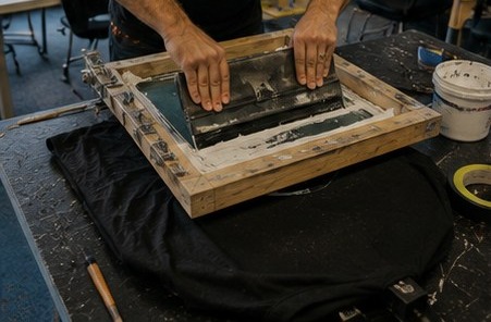 Hands pulling a squeegee across a silkscreen frame over a blank black t-shirt on a work table