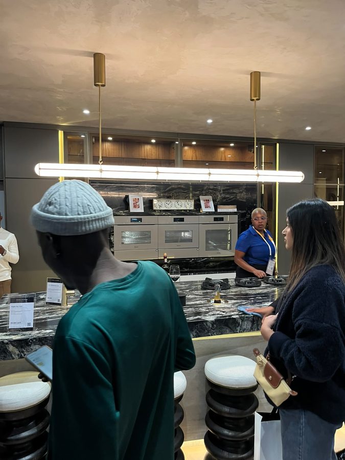 Kitchen showroom stand with stone counters, brass detailing and a linear pendant light, with visitors leaning on the counter